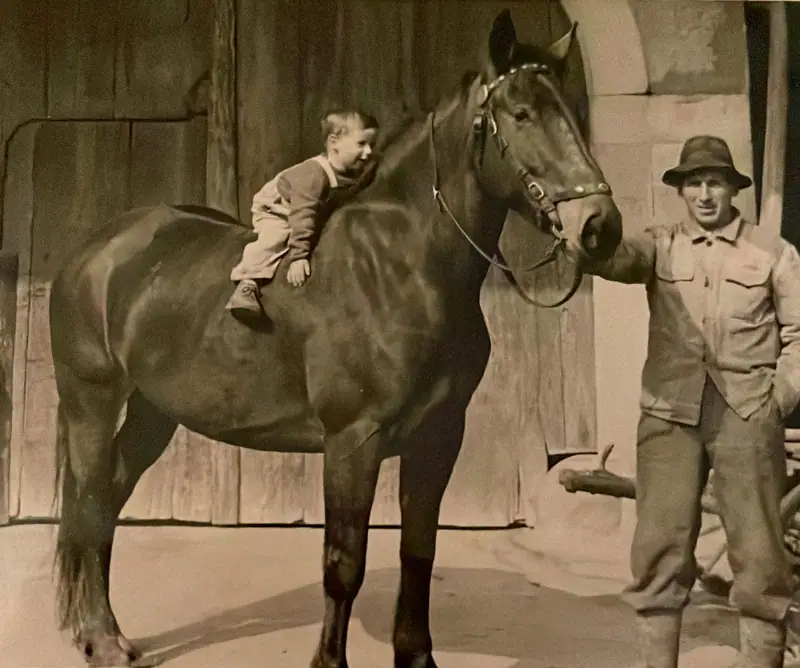 Historische Aufnahme: ein Kind sitzt auf einem Pferd, daneben steht ein Mann mit Hut vor einem Holztor
