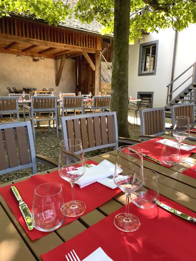 Rote Tischsets, Gläser und Besteck auf einem Holztisch im Innenhof, dahinter weitere Tische im Schatten