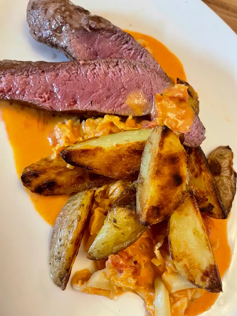 Rosa gebratene Fleischscheiben mit Kartoffelspalten auf orangefarbener Sauce