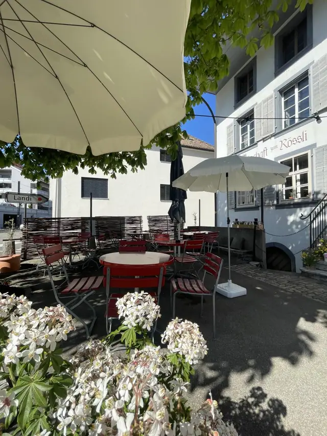 Die Terrasse zur Strasse: Tische, Stühle und Sonnenschirme, im Vordergrund weisse Blumen